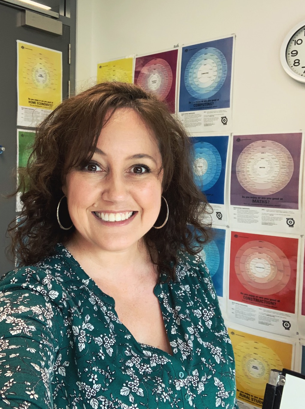Head shot of widely smiling woman, large round earrings, short, wavy dark hair, stands in front of wall with colourful posters.