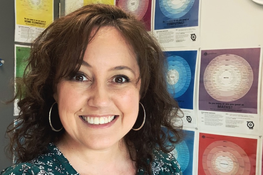 Head shot of widely smiling woman, large round earrings, short, wavy dark hair, stands in front of wall with colourful posters.