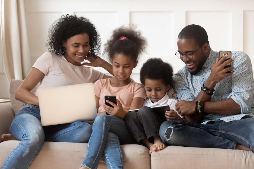 A black mother and father on the couch with their daughter and young son, guiding their use of a digital devices.