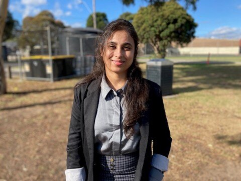 A woman wearing a jacket stands in a park and looks at the camera smiling.