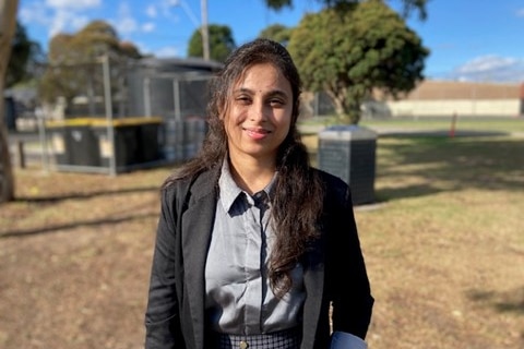 A woman wearing a jacket stands in a park and looks at the camera smiling.