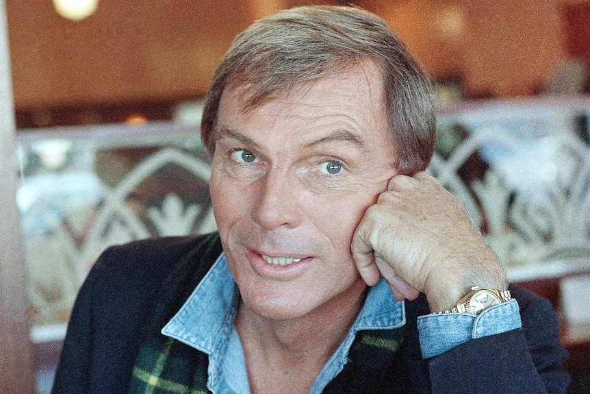 Adam West looks past the camera while seated at a table.
