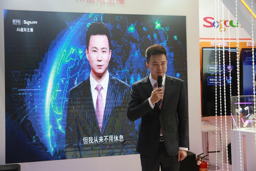 A man in a suit holds a microphone in front of a large poster with an image of an AI version of himself. 
