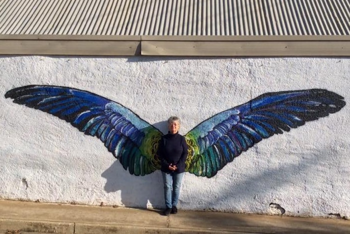 A woman is standing in front of a mural of blue and green wings, painted on a white wall.