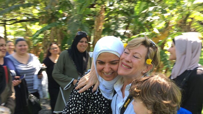 A woman in a hijab and a blonde woman holding a toddler, smile and embrace, with other women standing in the background