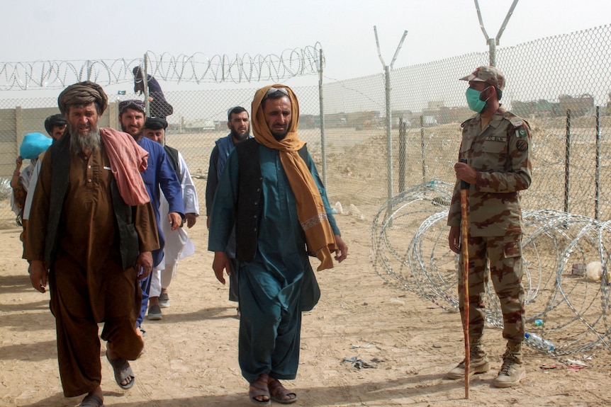 Middle Eastern men in traditional dress walk past a masked soldier next to a fence on a desert.