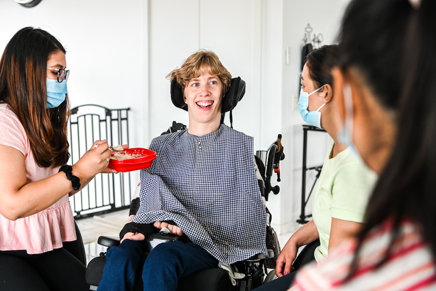 Rachel is in her wheelchair, being fed by carers. She has a beautiful smile and blue eyes.