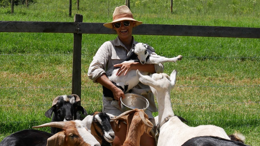 Goat farmer Cardia Forsyth holding a baby goat, surrounded by five pet goats on her farm