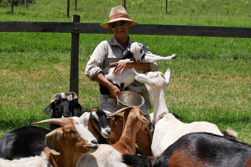 Goat farmer Cardia Forsyth holding a baby goat, surrounded by five pet goats on her farm