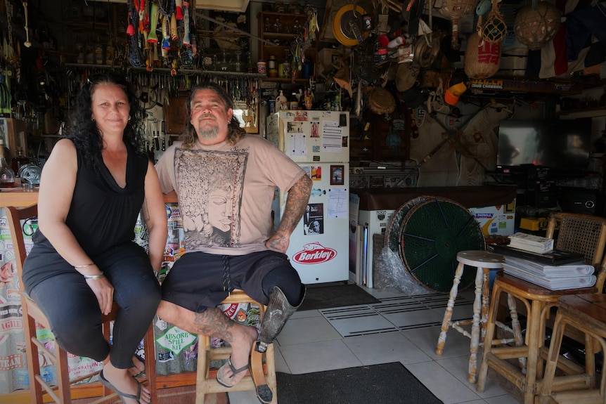 A smiling middle-aged woman and man sit in a shed on bar stools, firdge and lots of other stuff hanging from the roof.