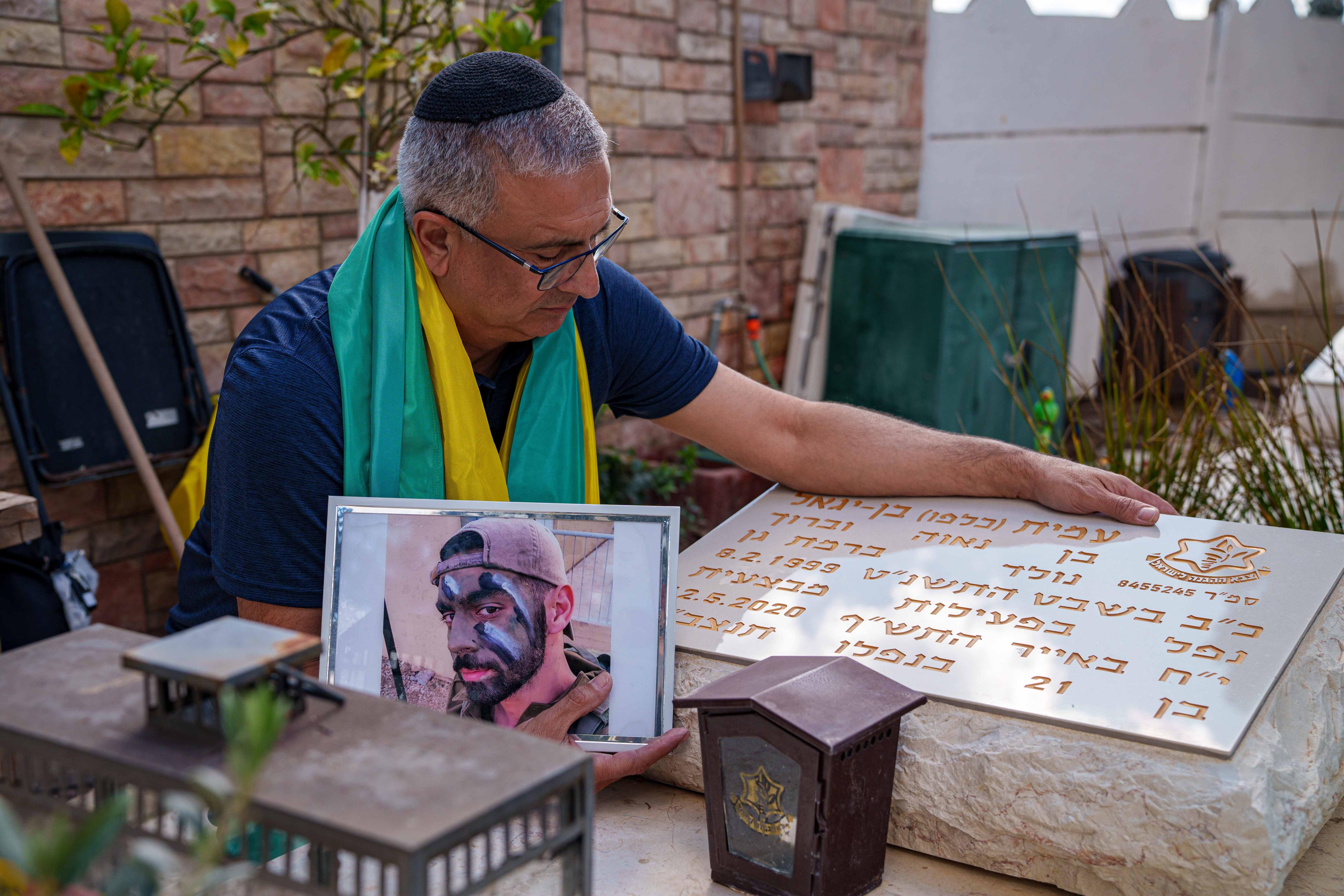 A man wearing a green and yellow shawl leans over a grave while holding a photo of his son.