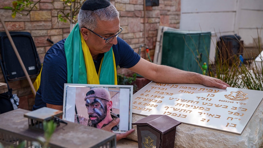 A man wearing a green and yellow shawl leans over a grave while holding a photo of his son.