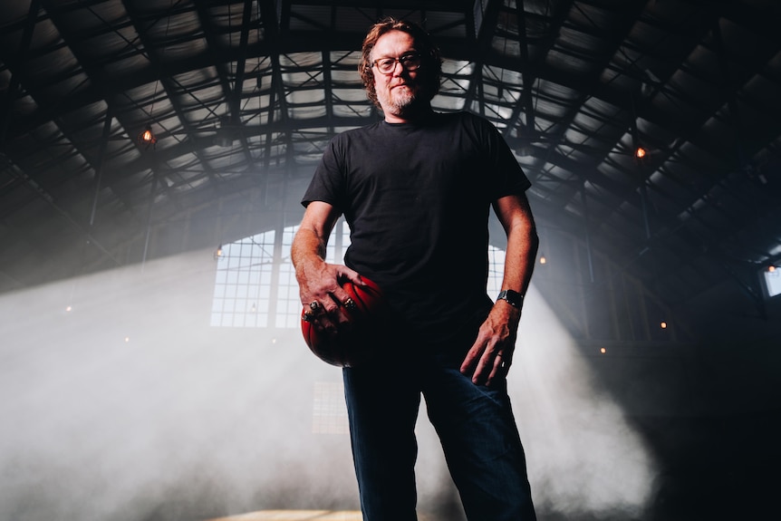 Luc Longley wears a black shirt and holds a basketball with window light streaming in behind him.