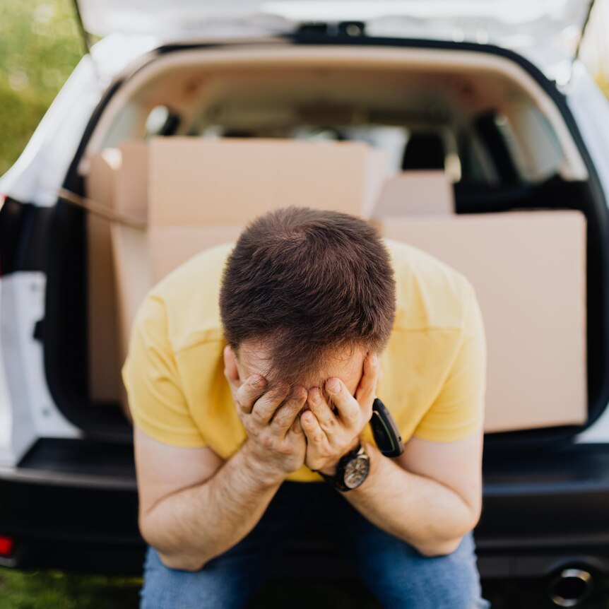 A man sitting on the back of a car with the boot open showing packing boxes in the car. He looks sad with his head in his hands