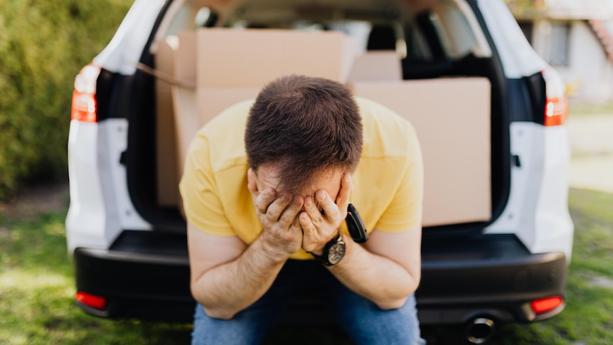 A man sitting on the back of a car with the boot open showing packing boxes in the car. He looks sad with his head in his hands