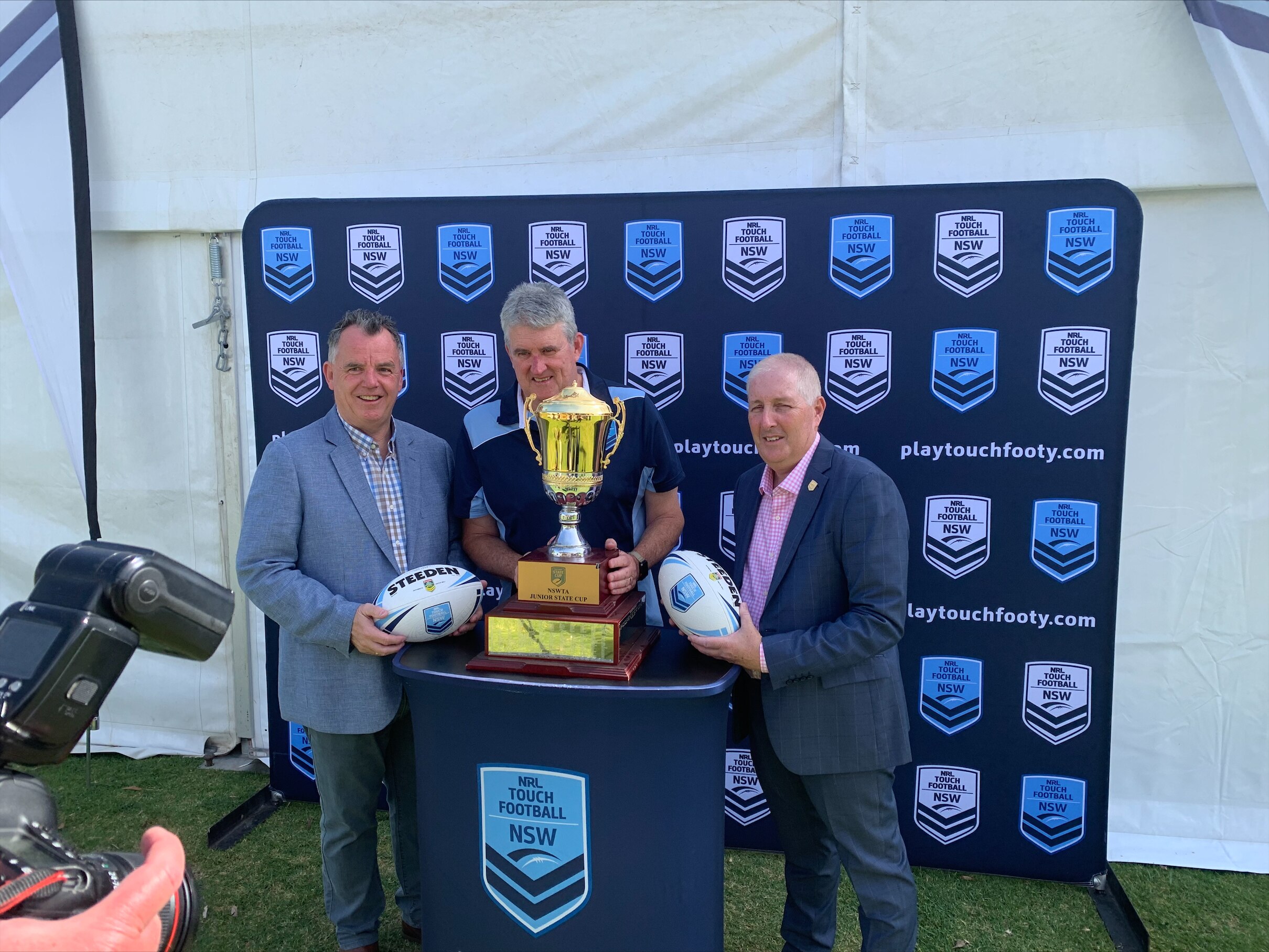 Three men in suits stand holding footballs and a trophy