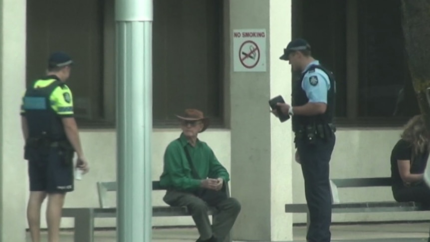 Police question a man sitting at a bus stop.