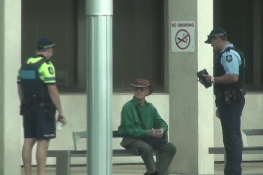 Police question a man sitting at a bus stop.