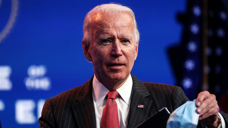 You see an elderly man in a pin-striped suit and red tie, holding up a disposable mask.