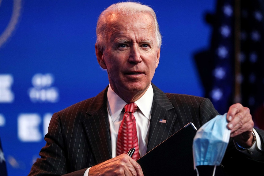 You see an elderly man in a pin-striped suit and red tie, holding up a disposable mask.