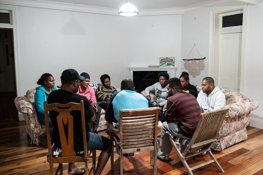 Nine people sit in a circle in a sparsely decorated loungeroom, some on sofas, others on outdoor chairs.