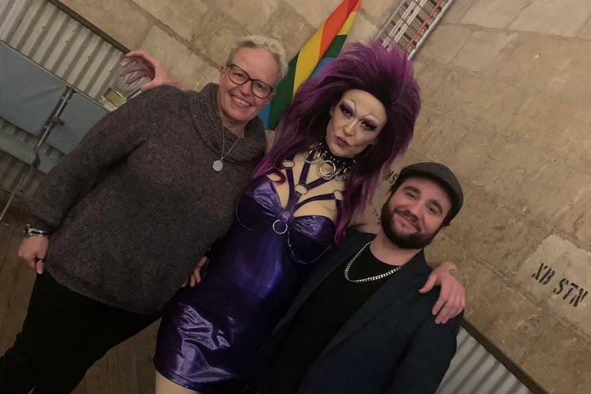 A man and a woman post with a drag queen in front of a rainbow flag. 
