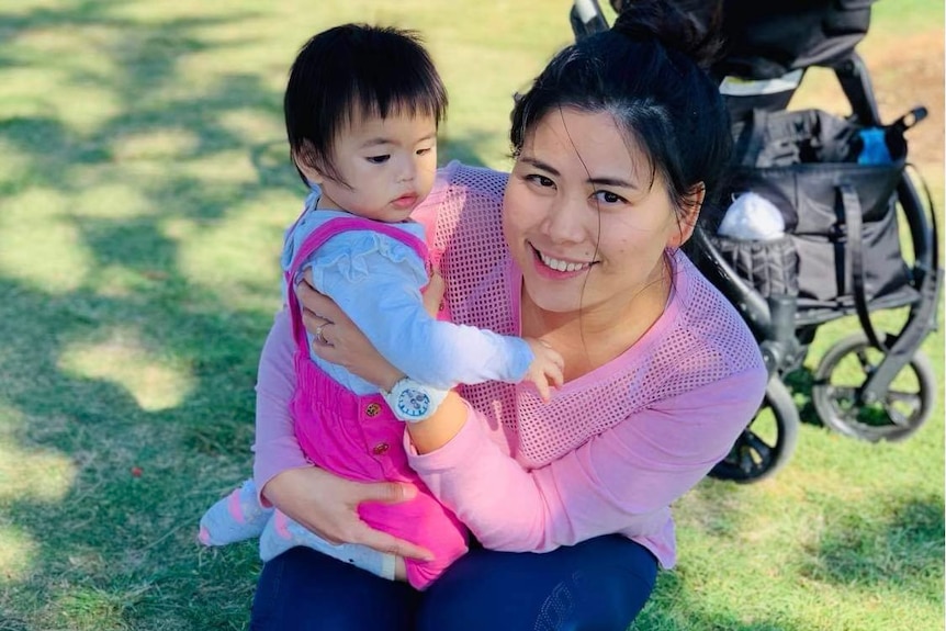 A mum in a pink jumper crouches down while holding her baby. She is smiling widely.