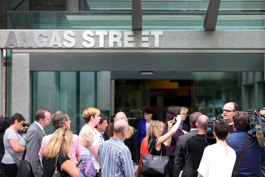 A crowd of people stand outside a court building, members of the media are filming while other people stand and watch.