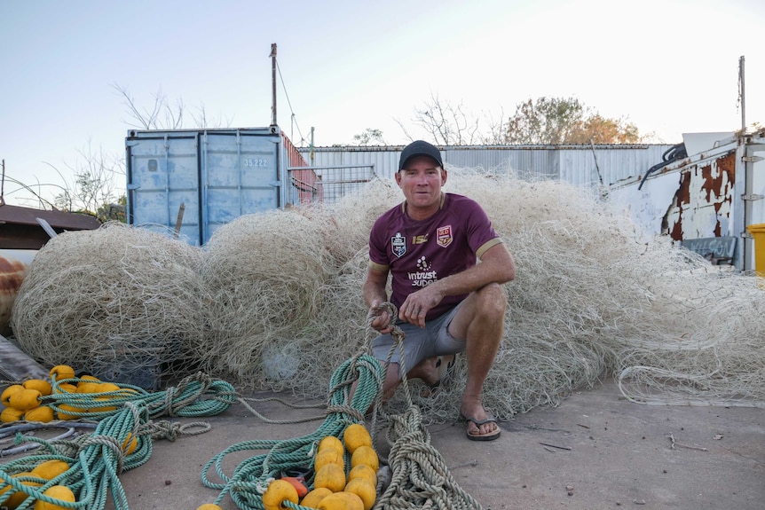 An image of a man kneeling before old fishing netting, wearing a Queensland maroons jersey.