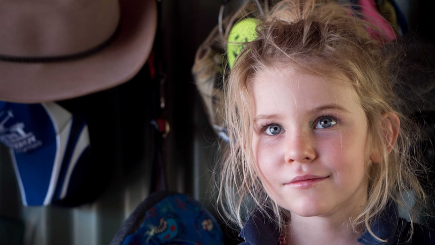 Young girl with blonde hair and blue eyes stares up at the camera in front of an outside wall with hats hanging on it.