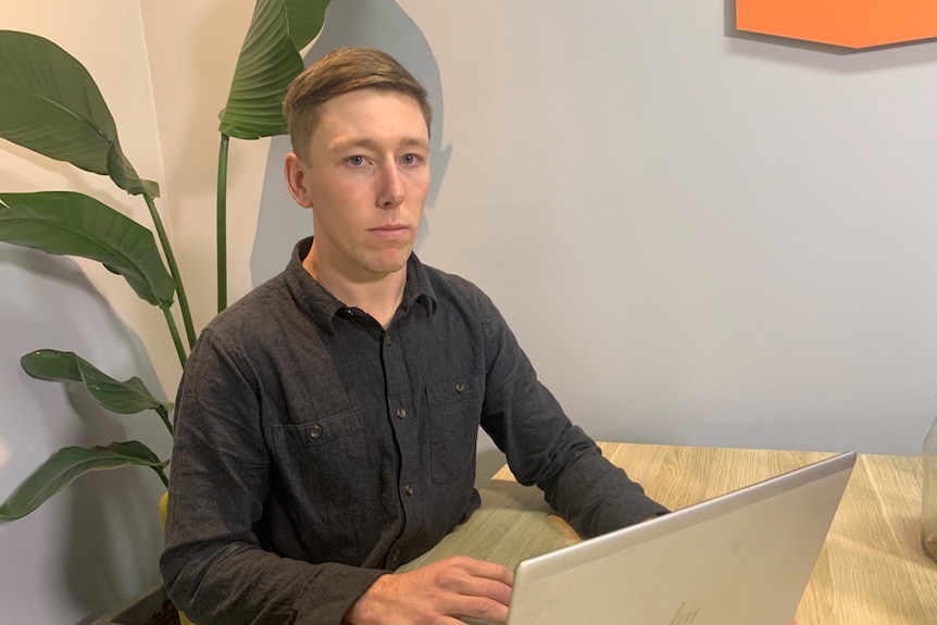 A serious young man with dark brown hair man sits at table in front of computer, a plant behind, an orange frame on the wall.