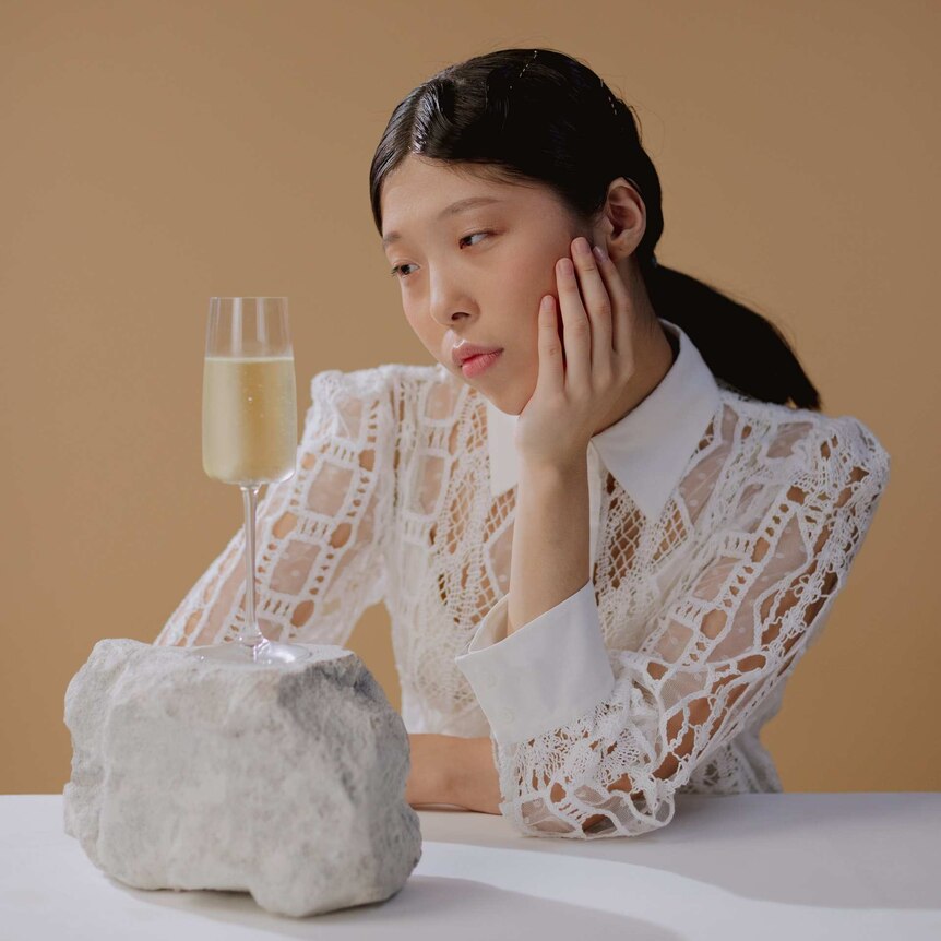 a woman in a white dress stares at a glass of champagne sitting on a rock