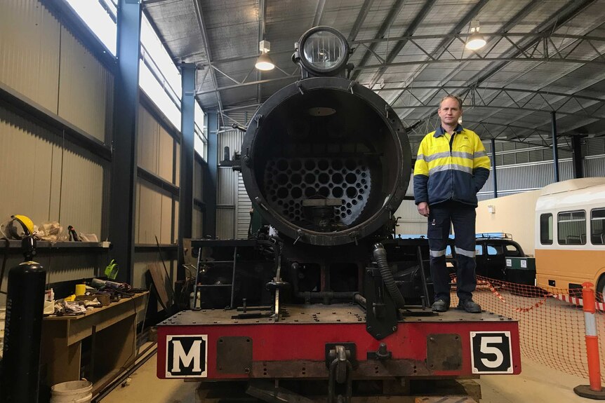 The front of a steam engine in a workshop with a man wearing high-vis looking down at the camera.