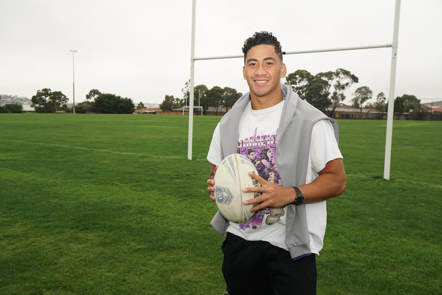 A  man in a Melbourne Storm t-shirt smiles as he holds a rugby ball on an oval.