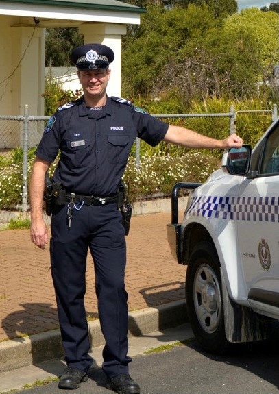 A smiling man in police uniform leans against a police car outside a police station