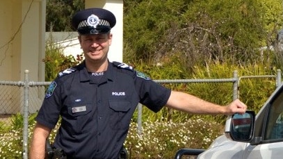 A smiling man in police uniform leans against a police car outside a police station