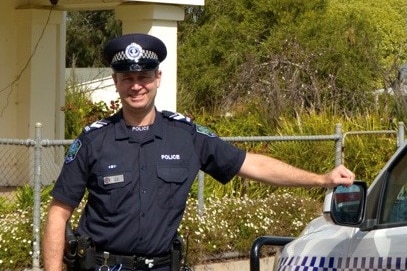 A smiling man in police uniform leans against a police car outside a police station