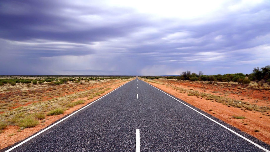 The highway stretches into the distance with red dirt and salt bush either side and ominous purple clouds above