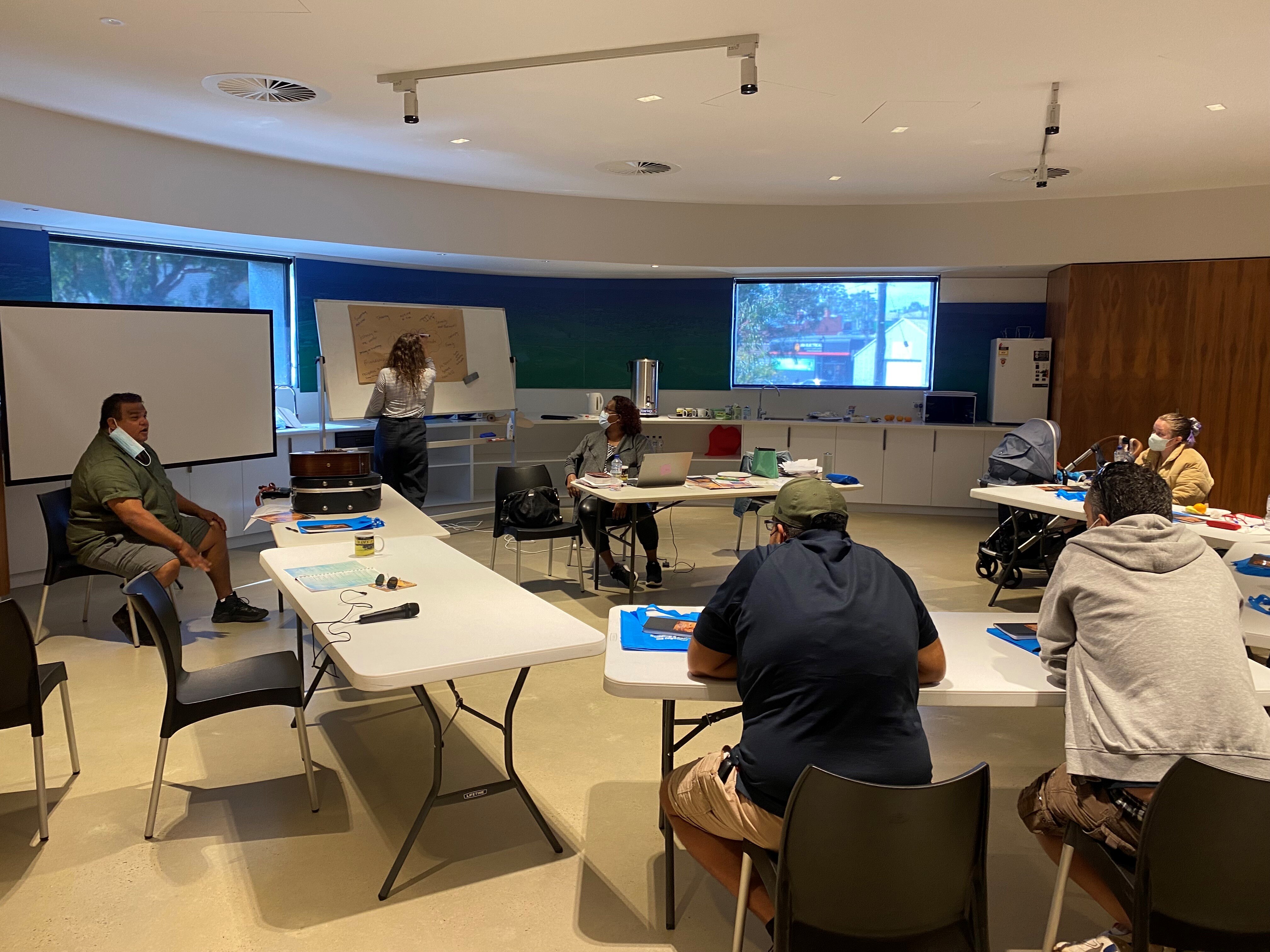 People sitting at desks in a classroom watch a woman write on a whiteboard