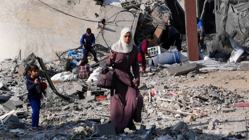 A woman in a headscarf carries bags through the rubble of a destroyed building