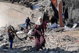A woman in a headscarf carries bags through the rubble of a destroyed building
