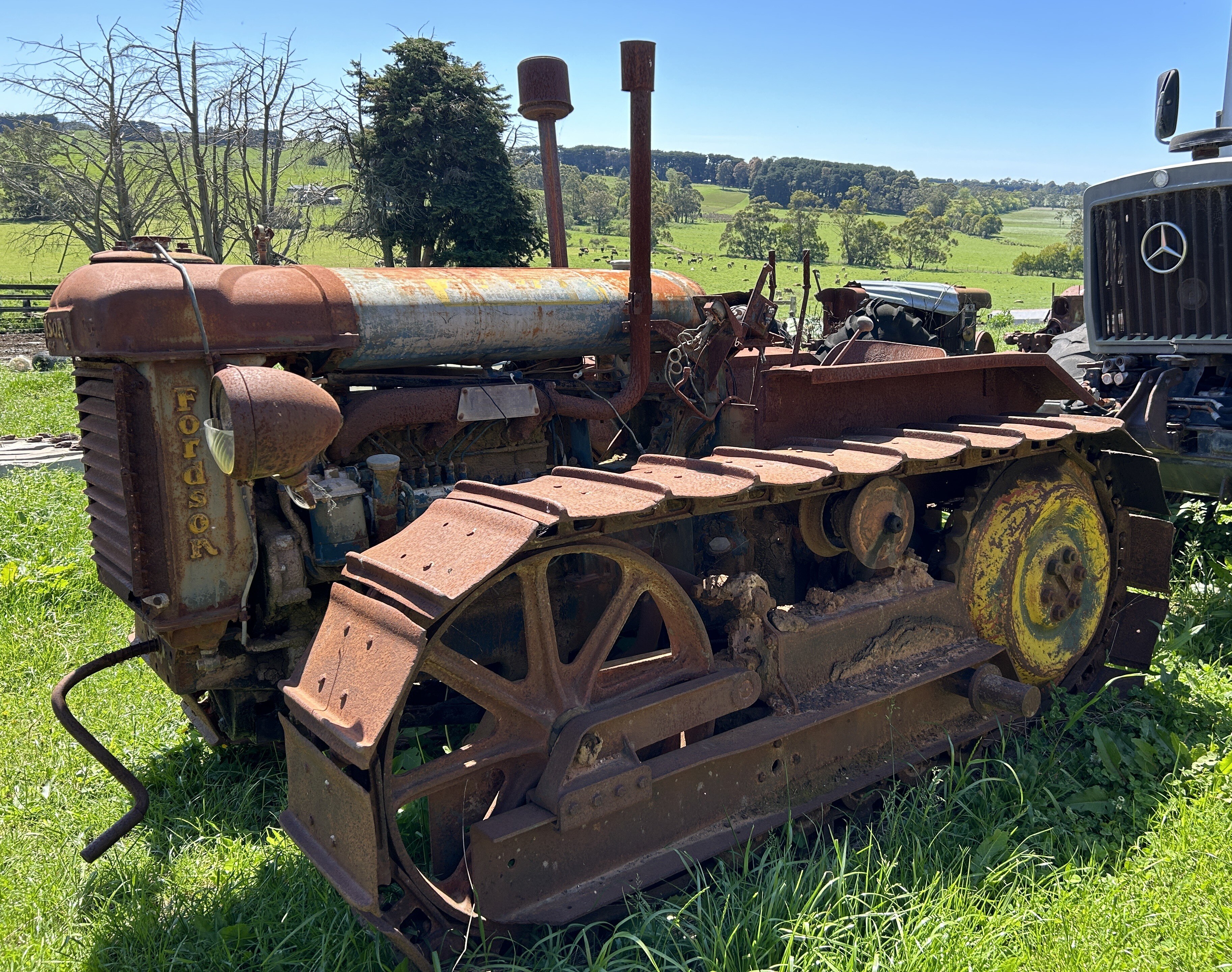 A rusty old tractor with caterpillar tracks sits in some grass on a country property.