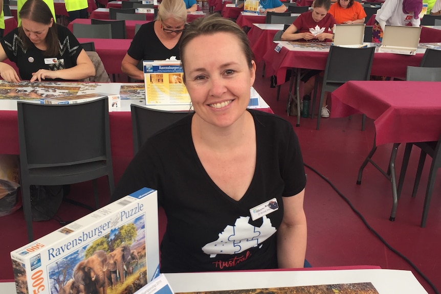 A woman smiles as she sits at a table with a completed jigsaw in front of her