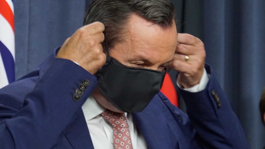 A mid-shot of WA Premier Mark McGowan putting on a black face mask after a media conference indoors.