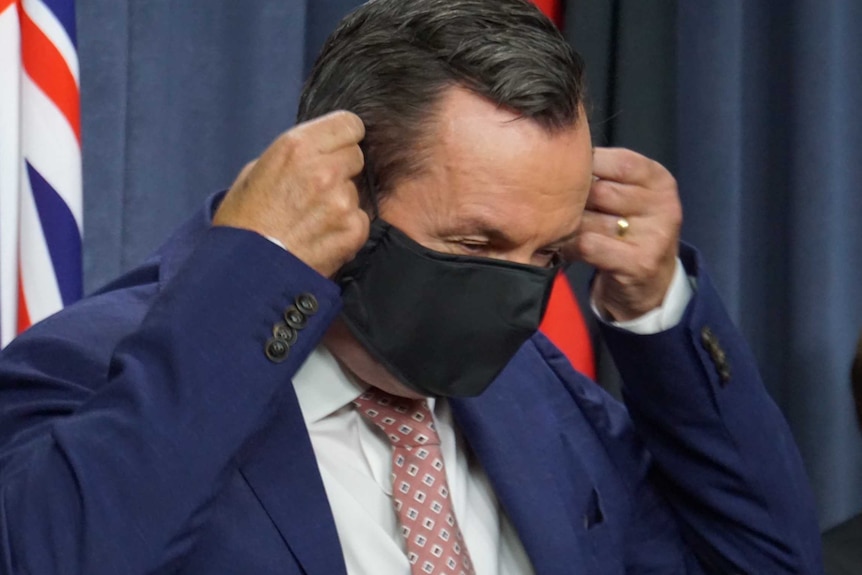 A mid-shot of WA Premier Mark McGowan putting on a black face mask after a media conference indoors.