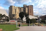 A gold-coloured building with other lower buildings and paving in front