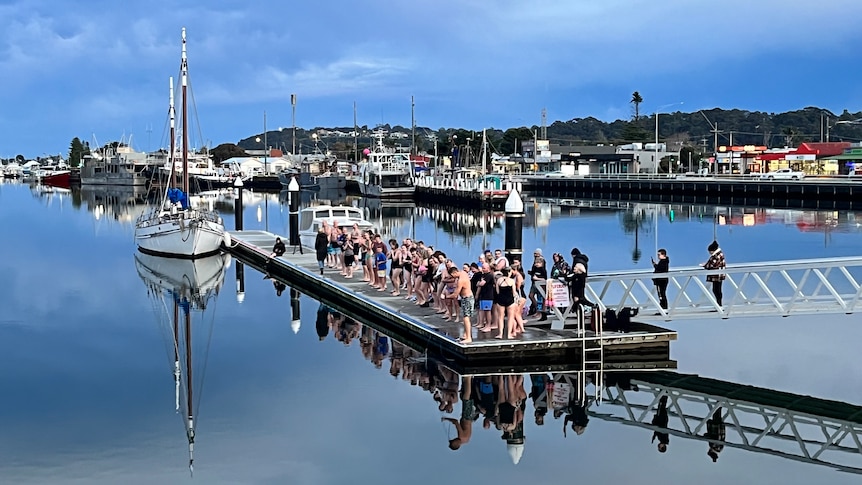 A group of swimmers standing on a jetty ready to jump into the water with boats in the background