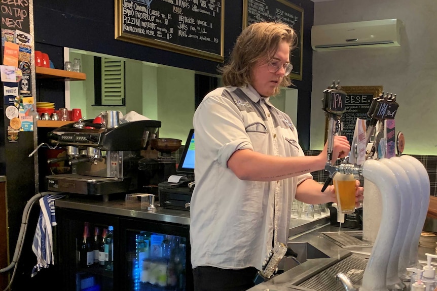 A blonde man wearing glasses pours a beer into a glass from a beer tap