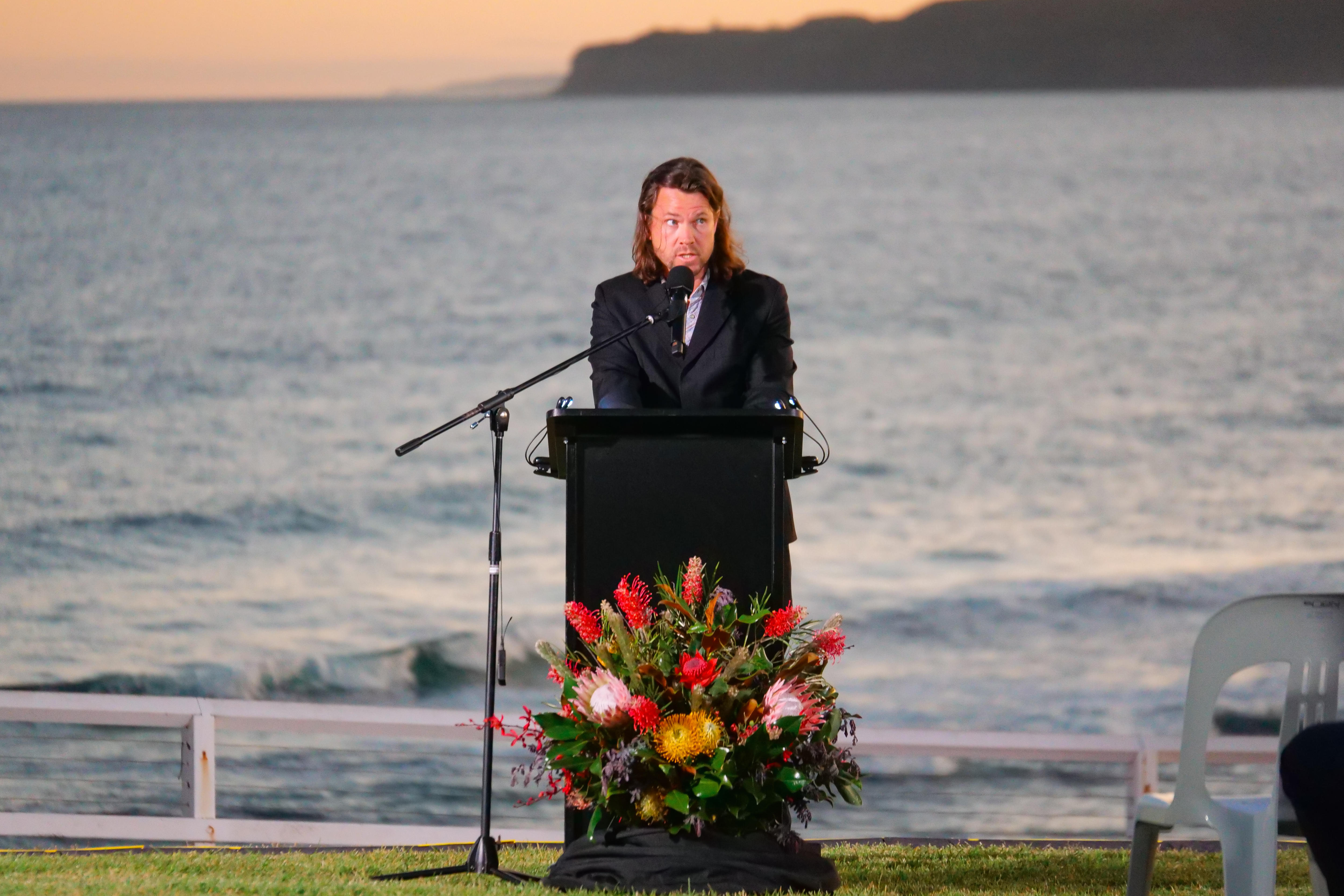 A man in a suit speaks at lectern with native flowers at the base outdoors, with ocean at sunset behind him.