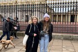 Two women wearing coats and a camera stand outside the gates of Buckingham Palace.